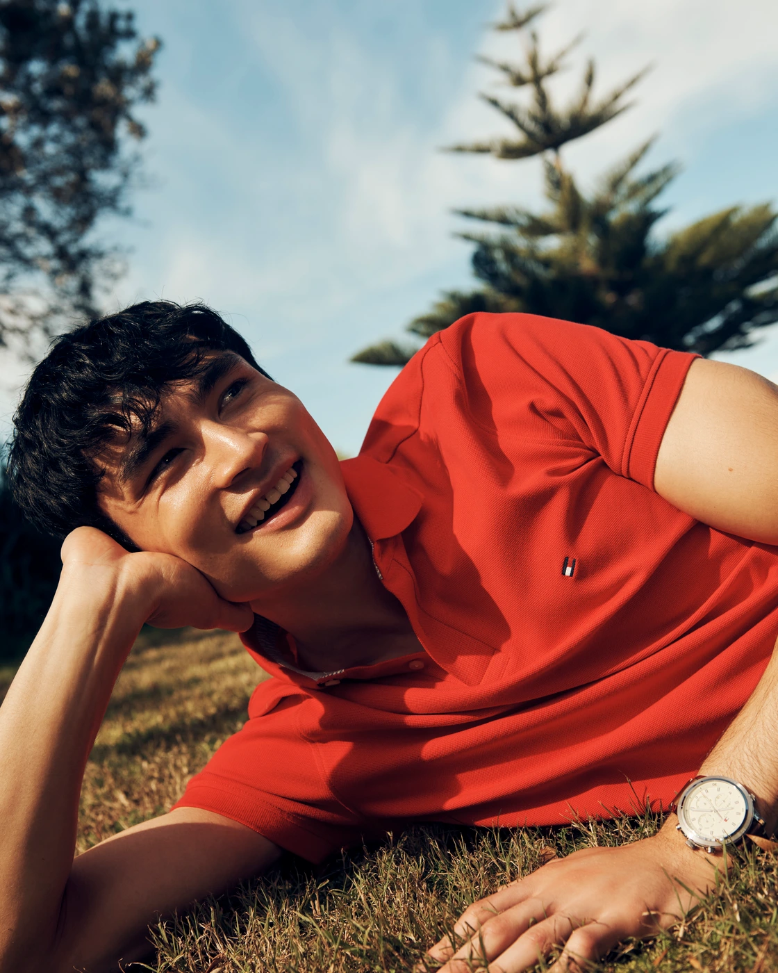 Man wearing a red polo shirt lying on grass under a clear blue sky, smiling outdoors