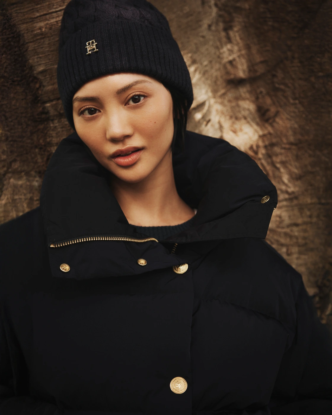 Close-up portrait of a woman wearing a navy puffer jacket and matching beanie standing against a natural wood tree trunk background