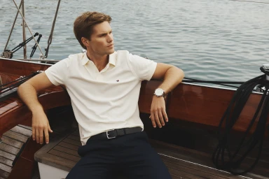 Man wearing a white polo shirt and dark trousers seated on a wooden boat deck against open water.
