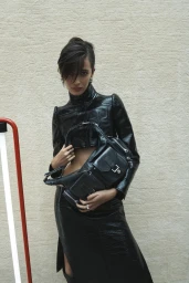 Model in black shiny jacket and skirt holding a black leather handbag against a textured wall