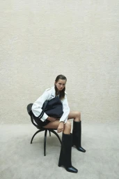 Model sitting in a black chair holding a dark bag while wearing a white jacket and tall black boots against a neutral textured wall.