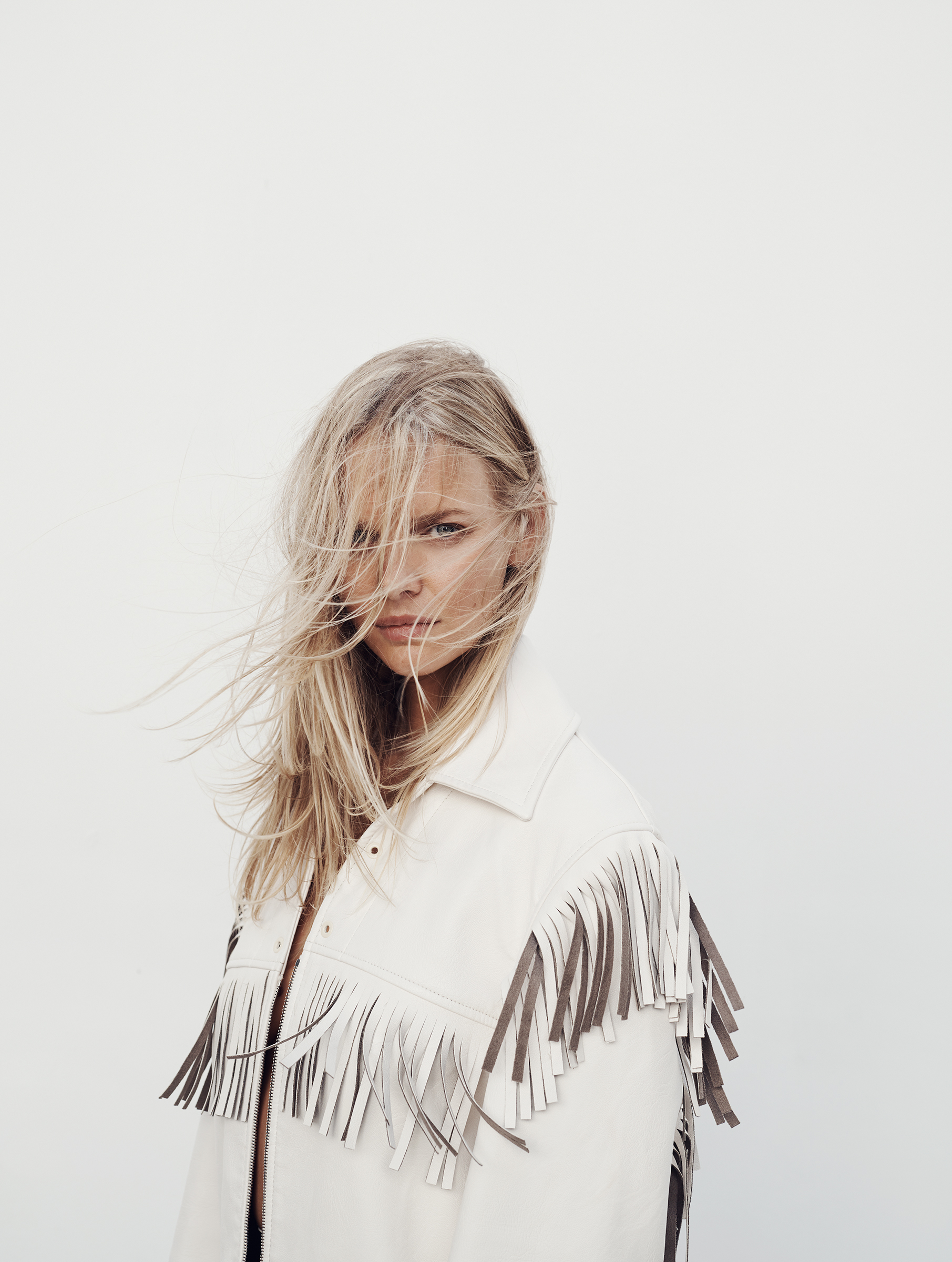 Close-up of a blonde woman with windblown hair, wearing a light jacket with fringes, set against a white background, fashion editorial style.