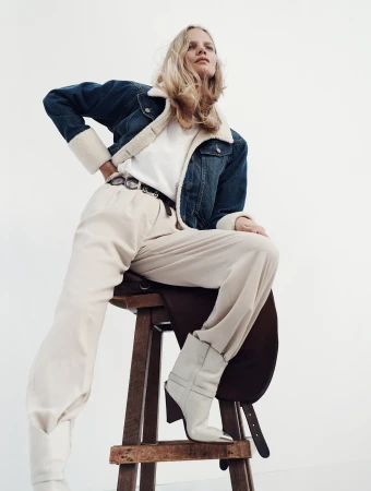 A blonde adult woman in a denim jacket and white pants sits on a stool in a studio, looking up and to the right in a fashion-style studio shot.