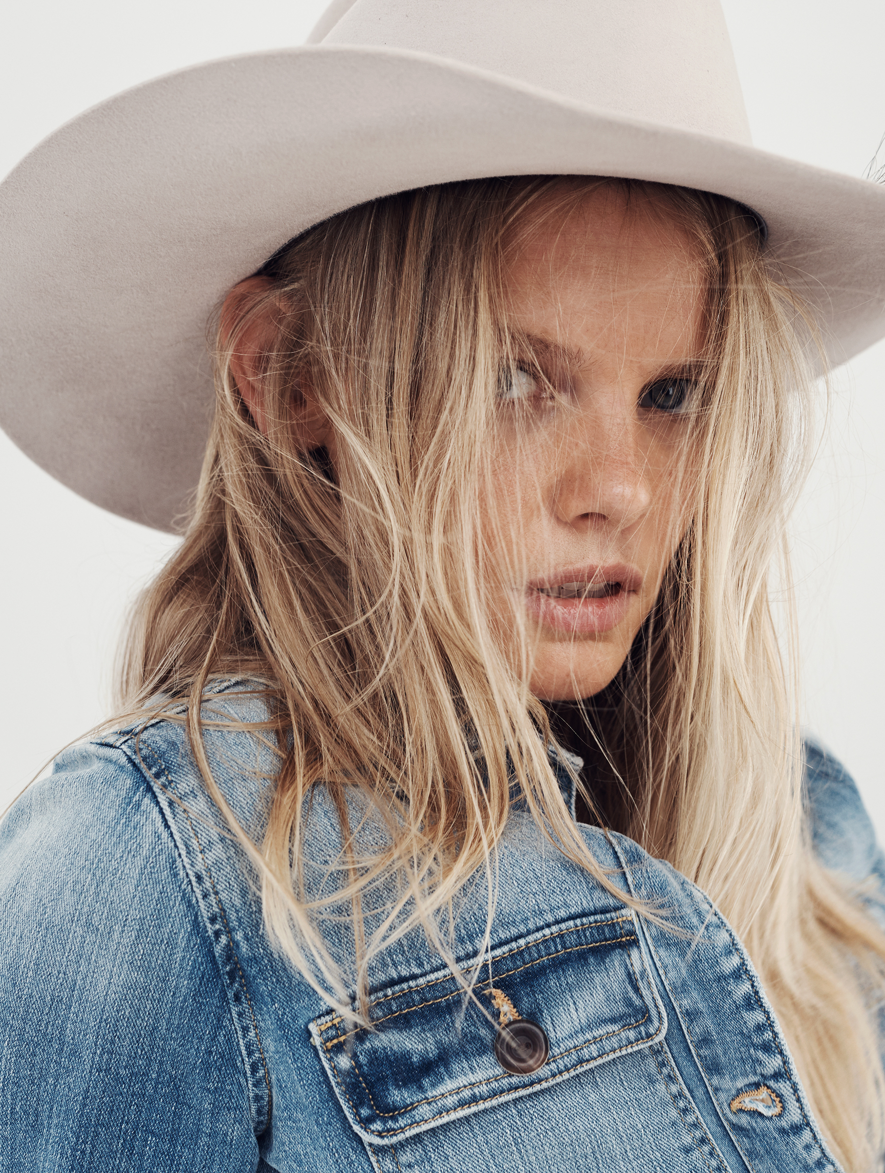 Portrait of a blonde woman in a denim jacket and a white cowboy hat against a neutral background, showcasing fashion and style.