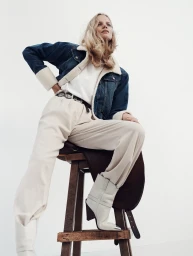 Model wearing shearling-trimmed denim jacket and white pants seated on a wooden stool against a white background
