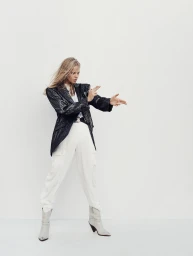 Model in a black blazer and white trousers posing with finger guns against a plain white studio background.