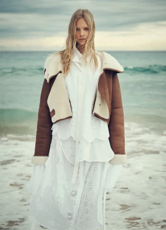 A blonde model in a white dress and shearling jacket stands on the beach with the ocean and cloudy sky as a backdrop.