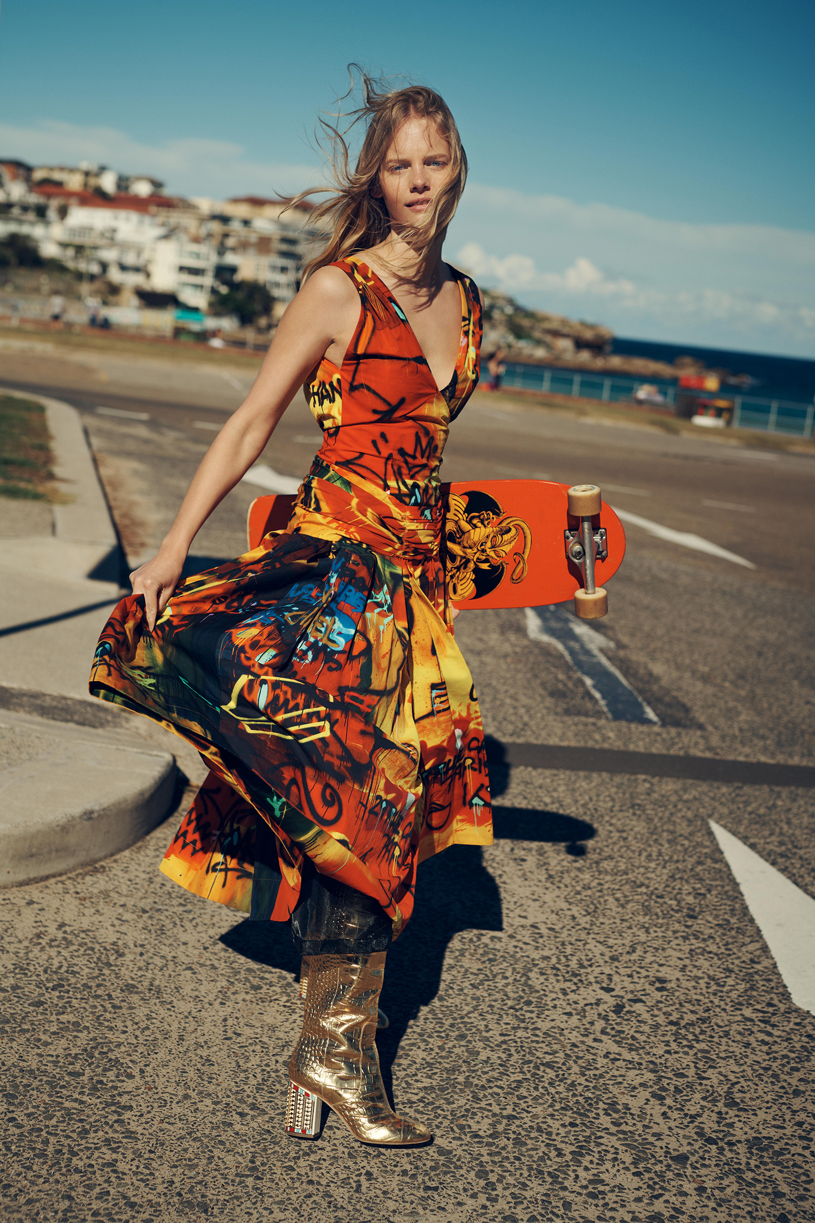 A blonde woman in a patterned dress and gold boots holds a skateboard on a cracked asphalt road, with a coastal town in the background.