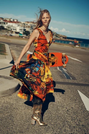 A blonde woman in a patterned dress and gold boots holds a skateboard on a cracked asphalt road, with a coastal town in the background.