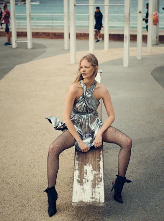 A woman in a metallic dress and black tights sits on an outdoor bench near the beach in a fashion editorial shot with diffused lighting.