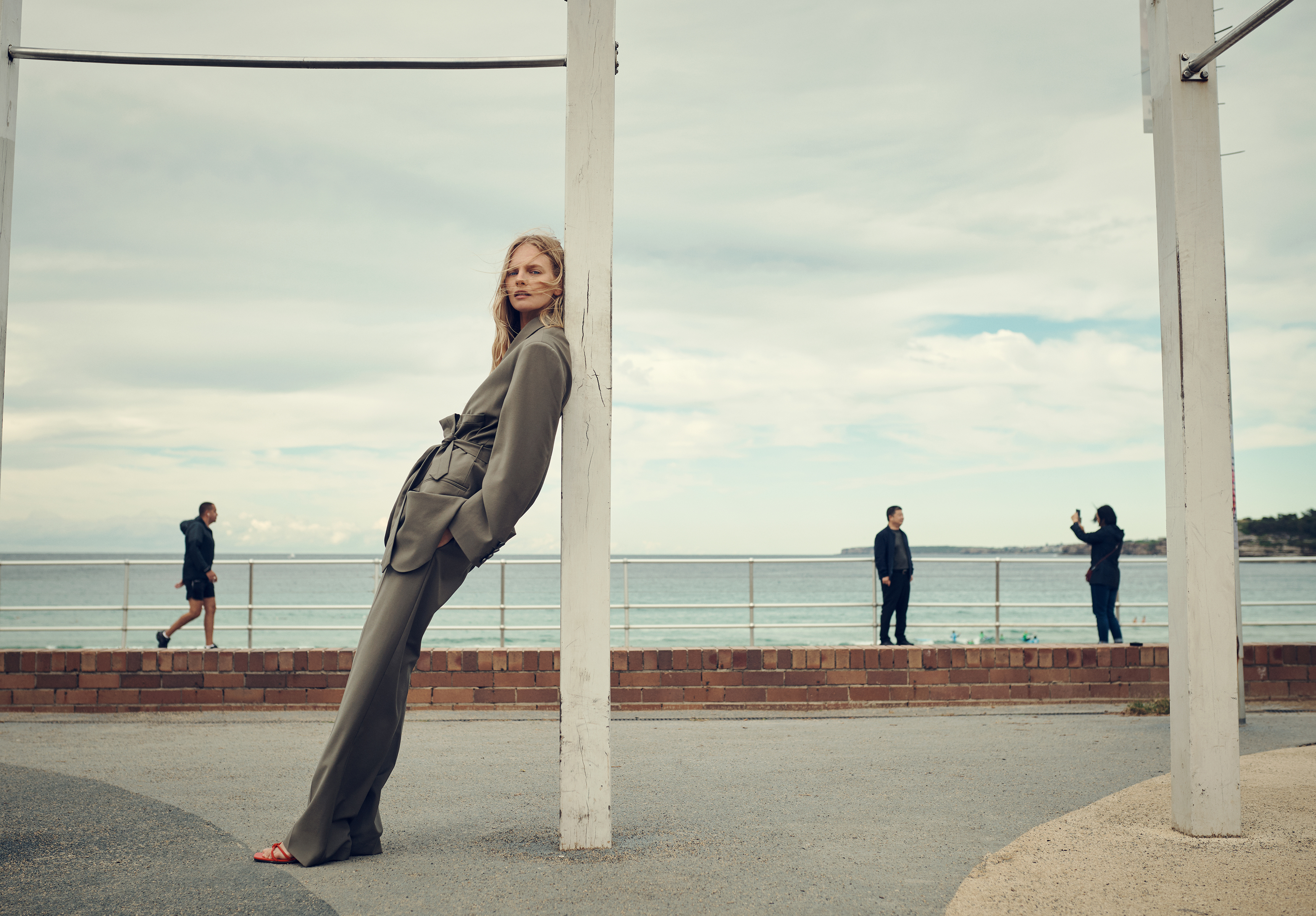 Fashionable woman in olive suit leans against post near ocean, featuring a cloudy sky, people in background and an urban setting.