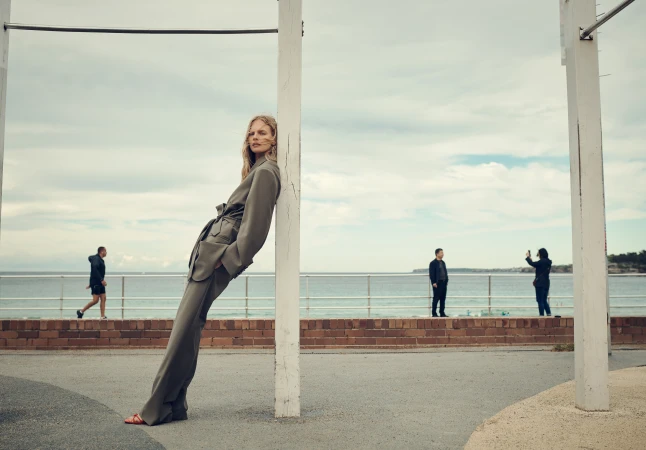 Fashionable woman in olive suit leans against post near ocean, featuring a cloudy sky, people in background and an urban setting.