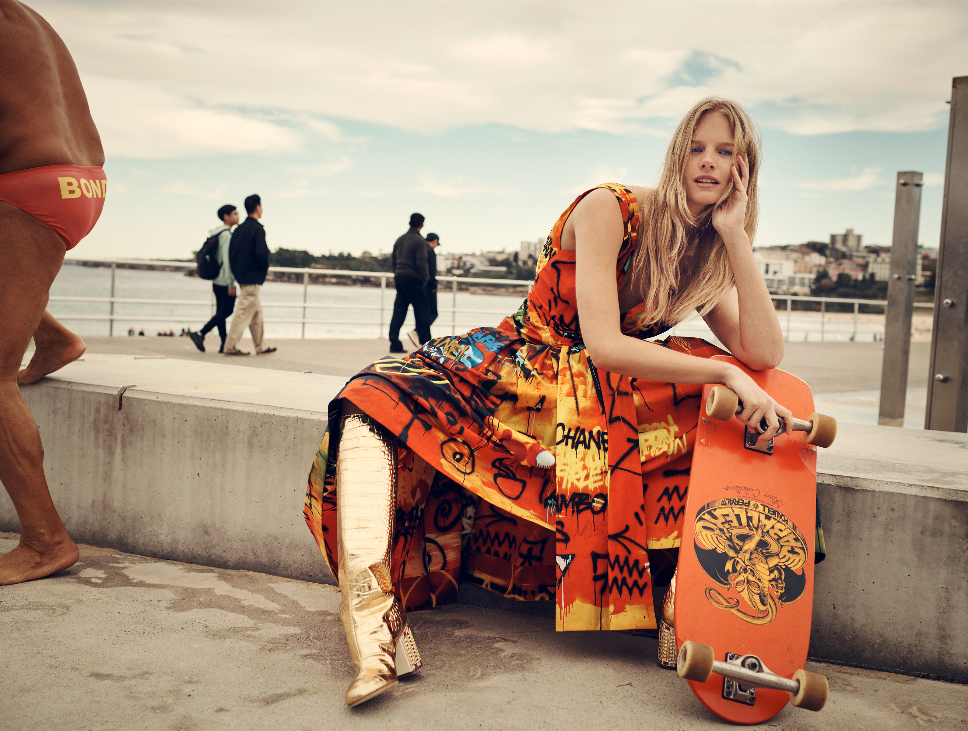 Woman in an orange dress and metallic gold boots sits on a concrete wall with a skateboard on a beach promenade on a sunny day.