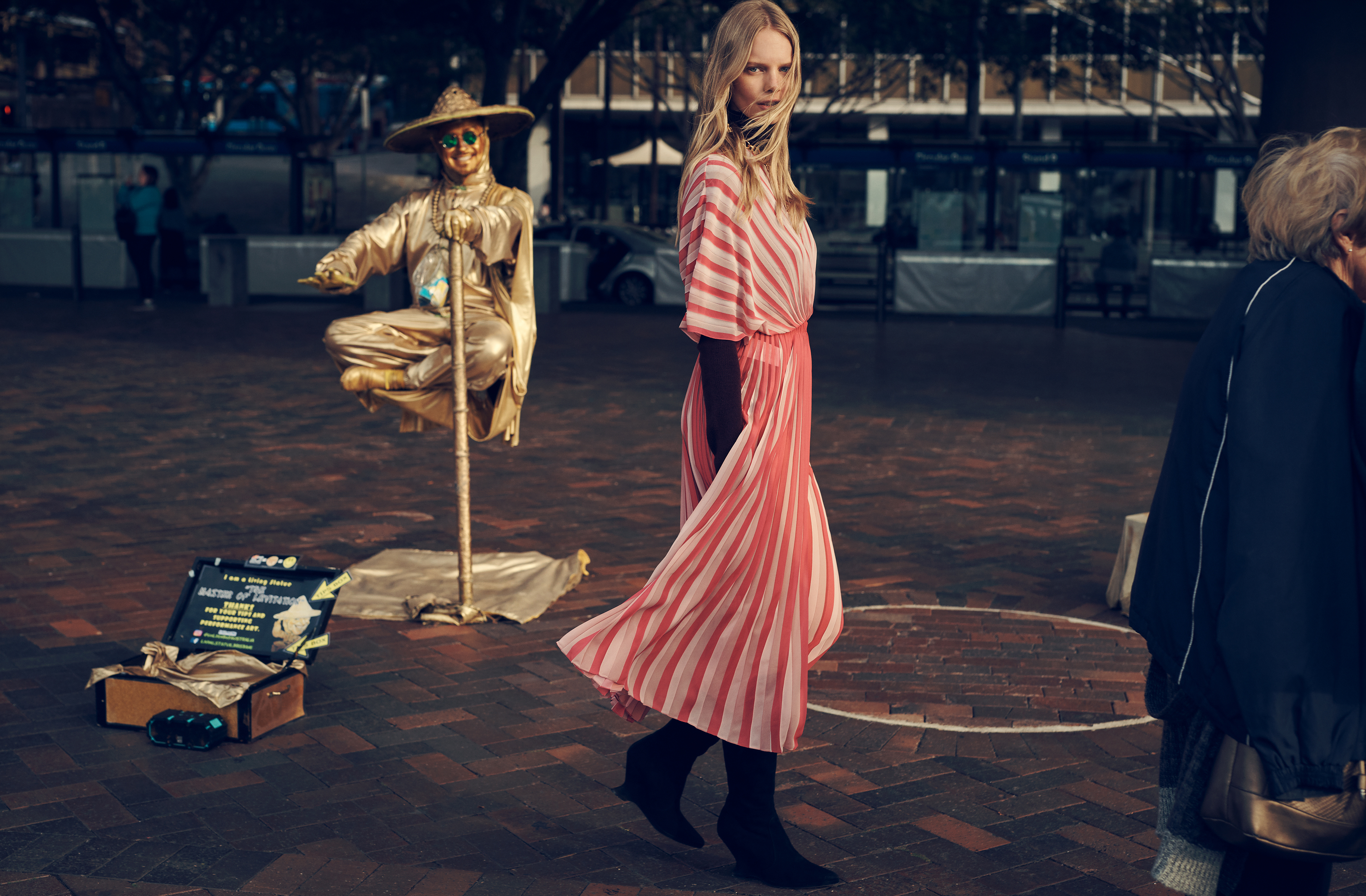 A woman walks past a golden street performer in an outdoor plaza. The performer sits cross-legged atop a support pole.