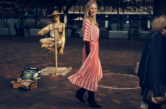 A woman walks past a golden street performer in an outdoor plaza. The performer sits cross-legged atop a support pole.