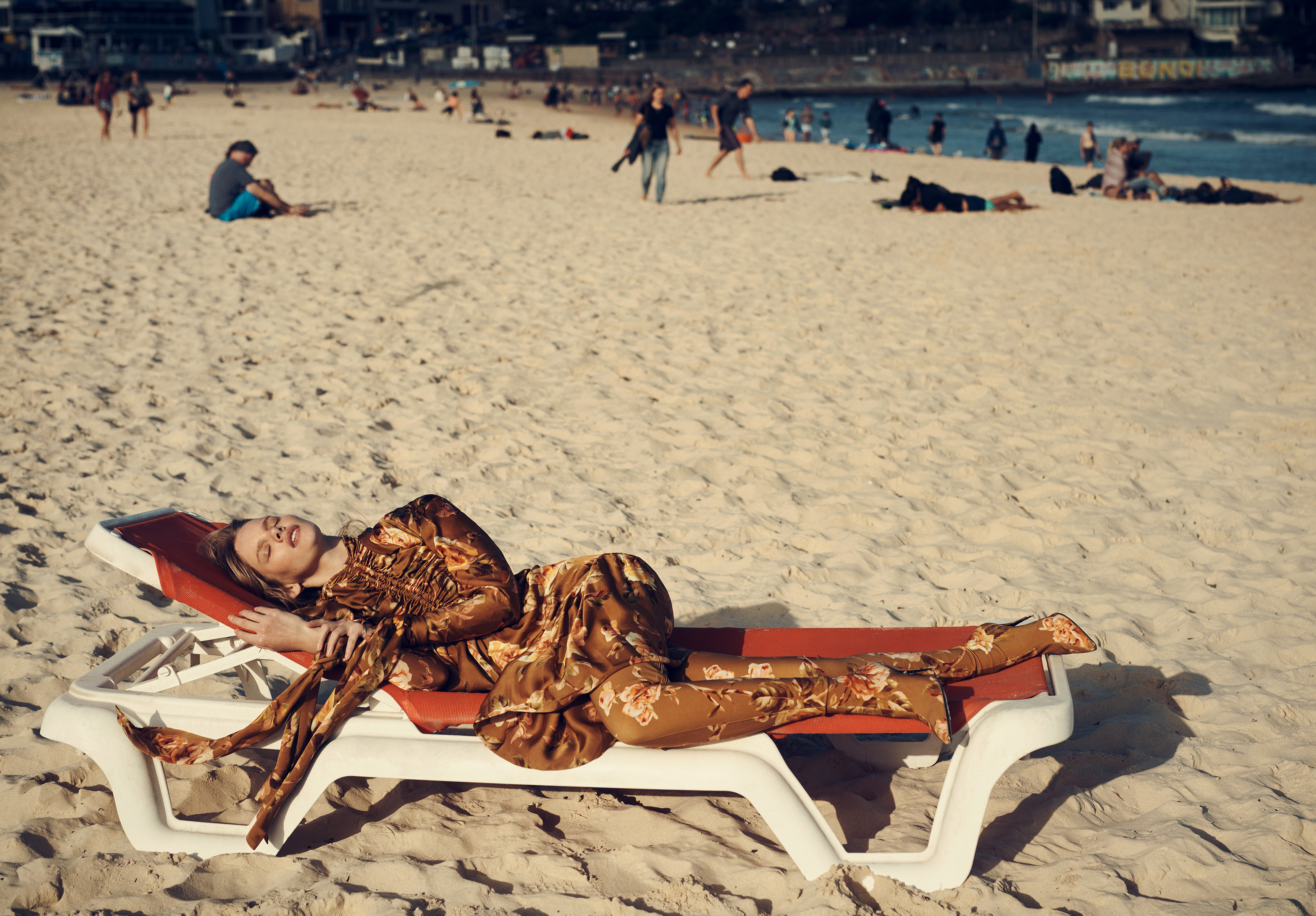 Woman in floral dress reclines on lounge chair on a sandy beach with other beachgoers and ocean visible in the background on a sunny day.