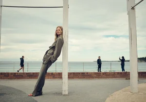 Model in grey trousers and blazer leaning against a white post by the ocean