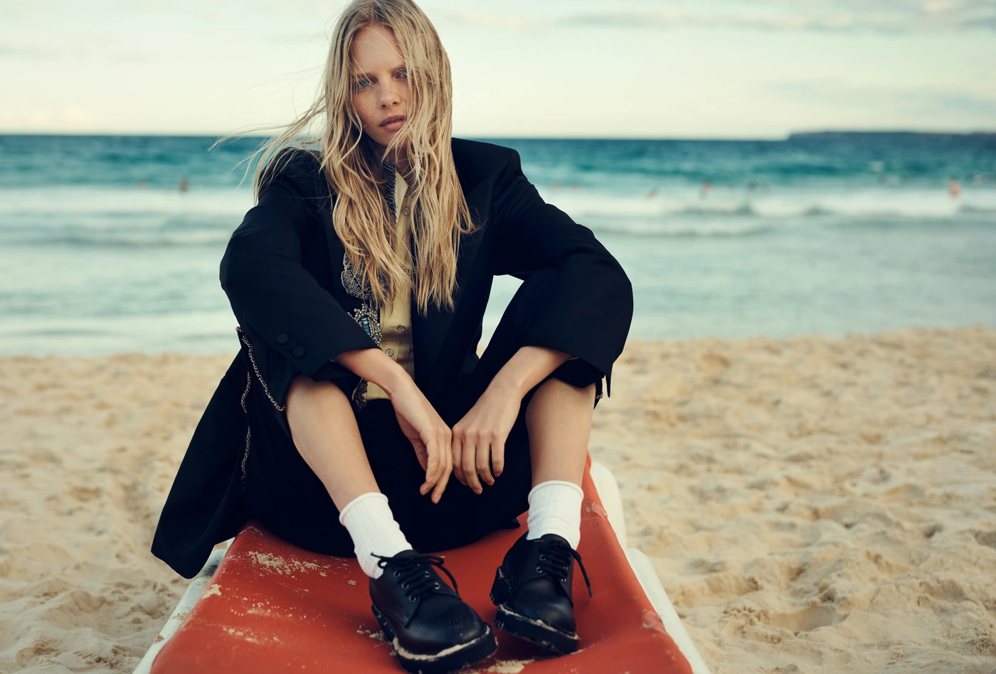 Model with long blonde hair seated on a beach wearing a black blazer and white socks with black shoes for Elle Australia.