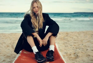 Model with long blonde hair seated on a beach wearing a black blazer and white socks with black shoes for Elle Australia.