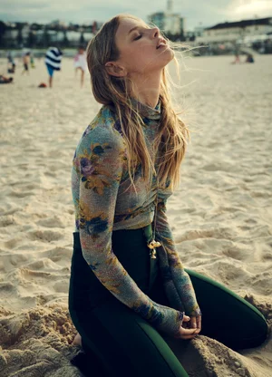 Model kneeling on a sandy beach wearing a floral turtleneck top and dark green trousers, photographed outdoors.