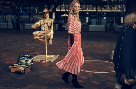 Model in pink striped dress walking on paved public square past a gold street performer for Elle Australia editorial