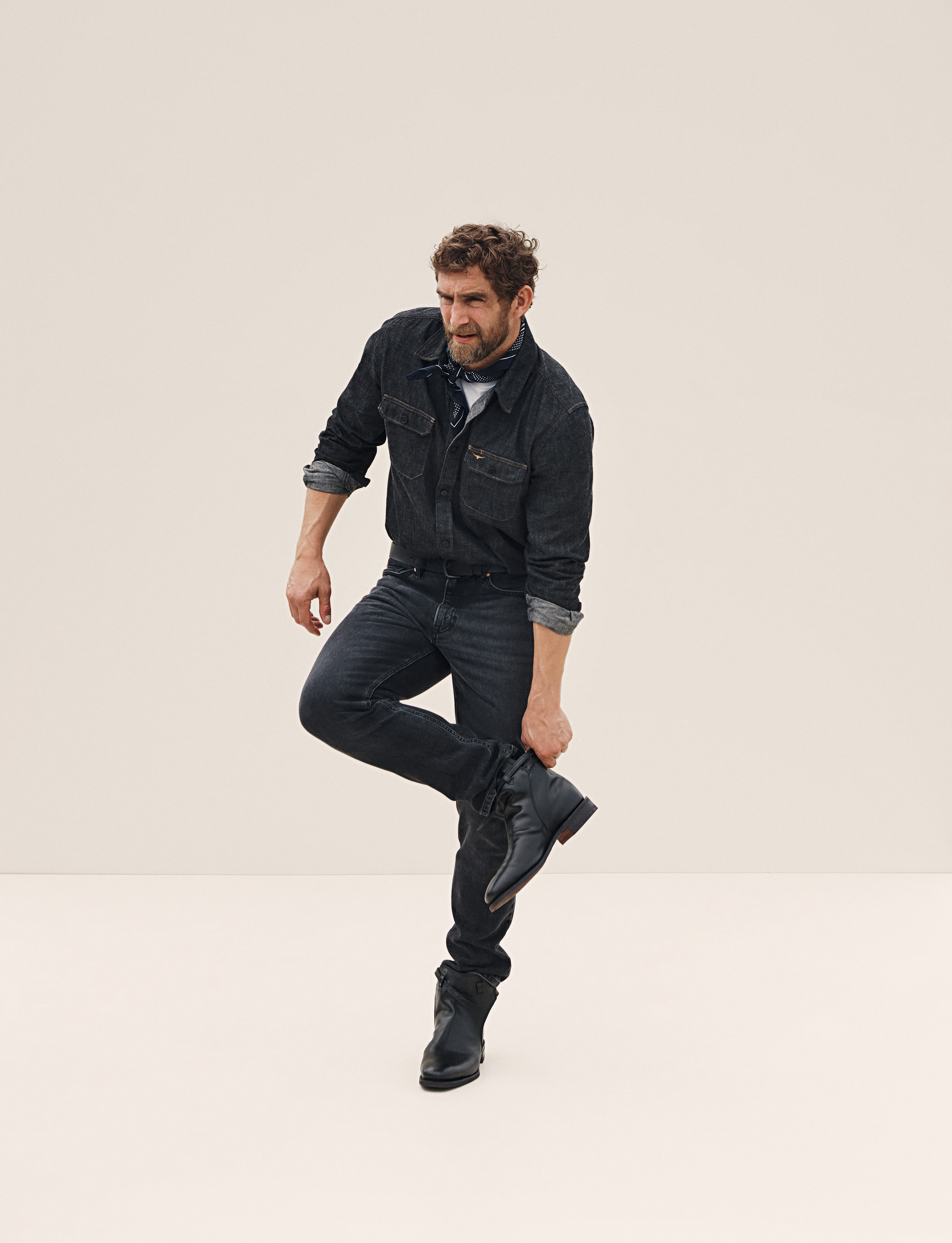 A man in a denim outfit balances on one leg, holding his boot in front of a plain background. He has a beard and is looking away.