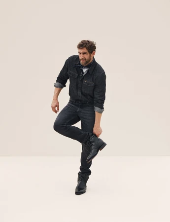 A man in a denim outfit balances on one leg, holding his boot in front of a plain background. He has a beard and is looking away.