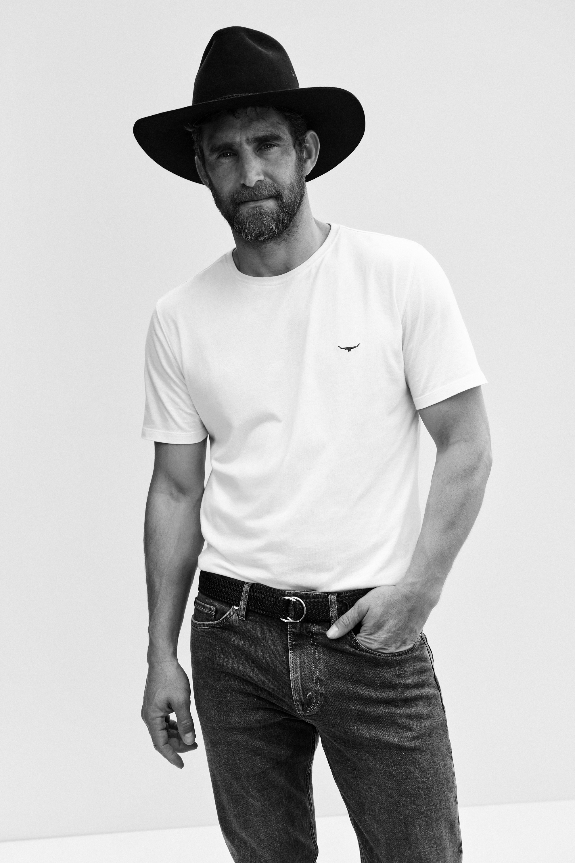 Monochrome portrait of a bearded man wearing a hat, white t-shirt, and jeans against a white background, posing with hand in pocket.