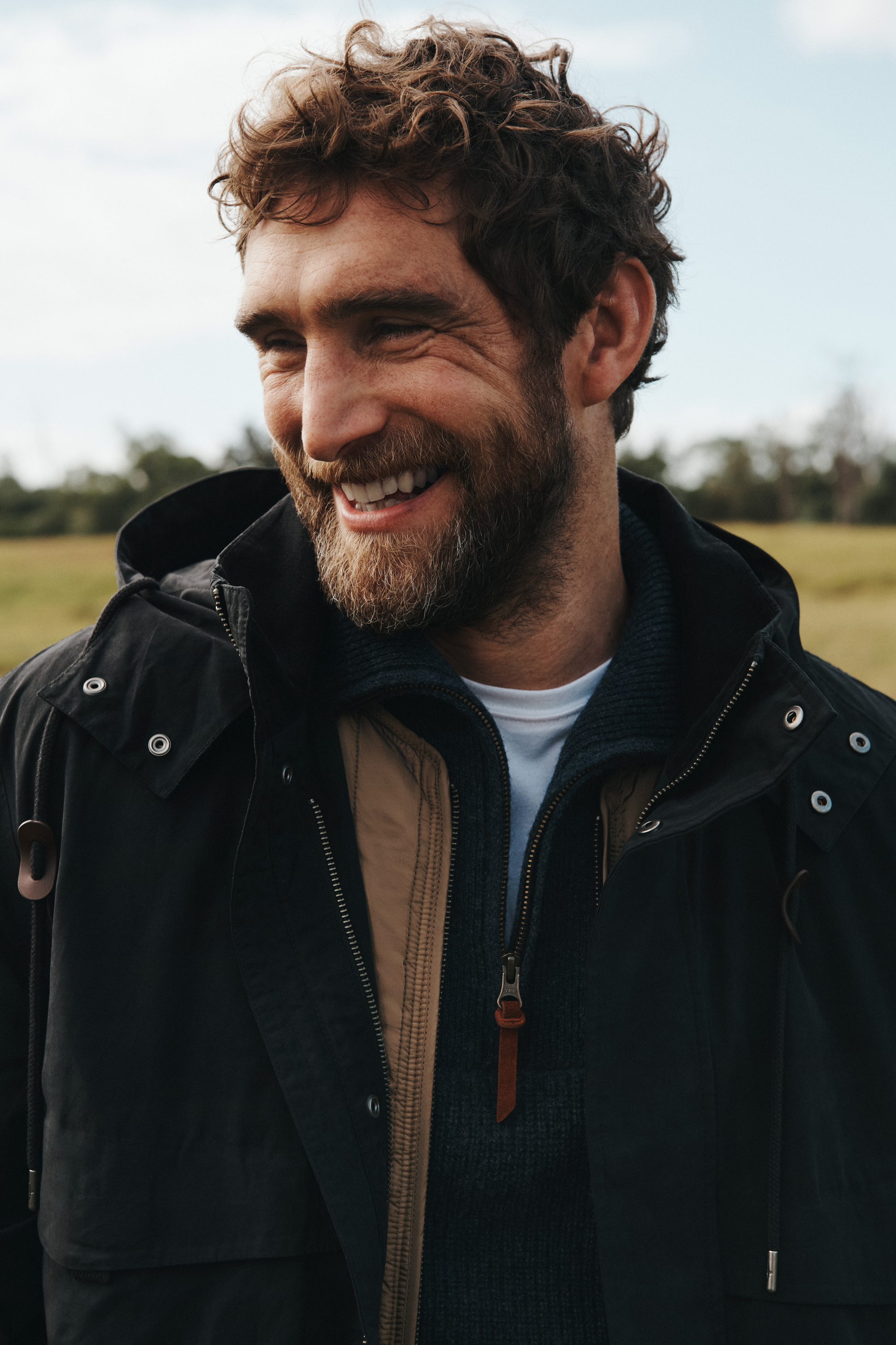 Man smiling outdoors in a dark coat with a beard and curly hair, standing in a grassy field with a blue and white sky above.