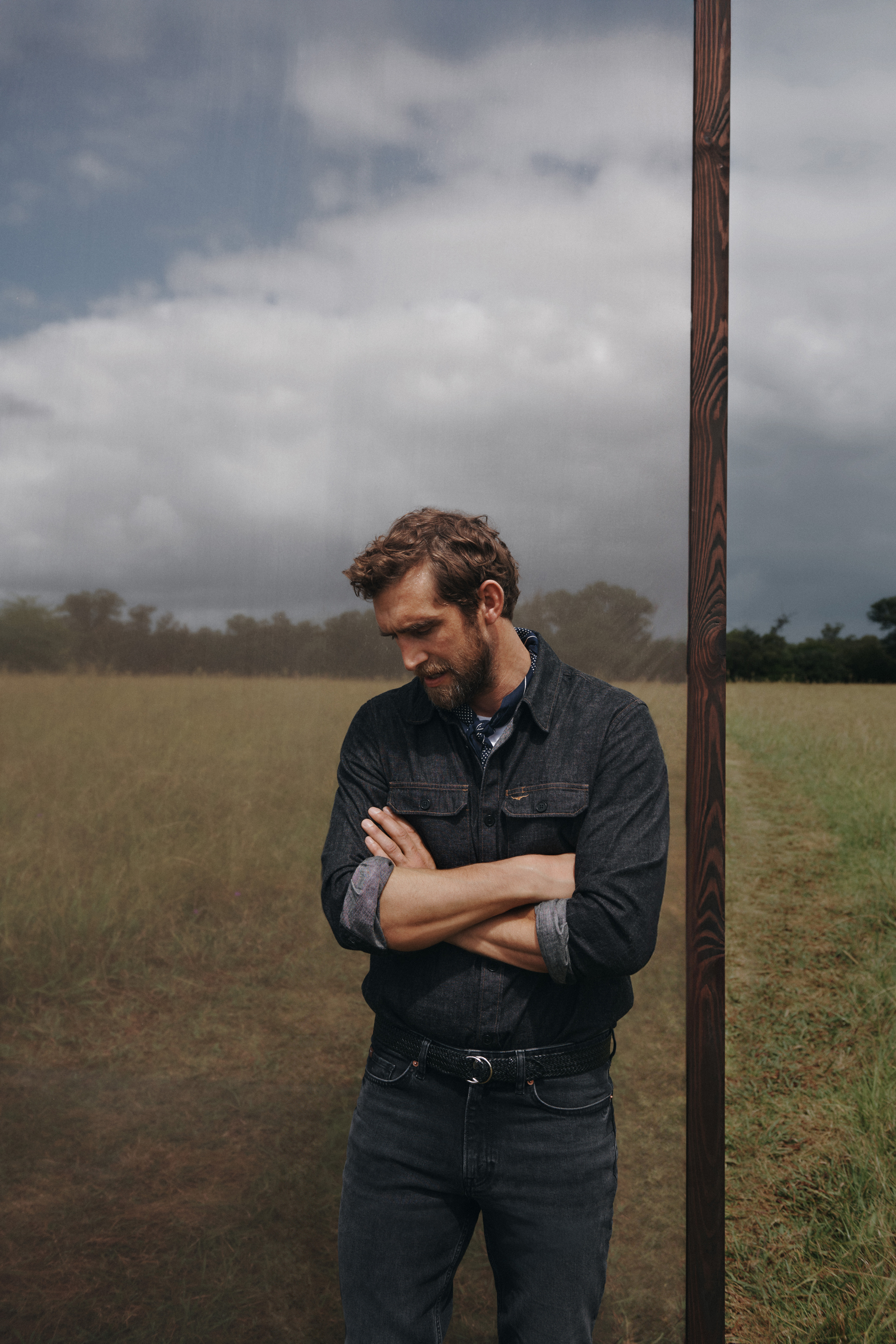 A man standing in a field wearing a denim shirt and jeans with his arms crossed, with a cloudy sky overhead. He is posing.