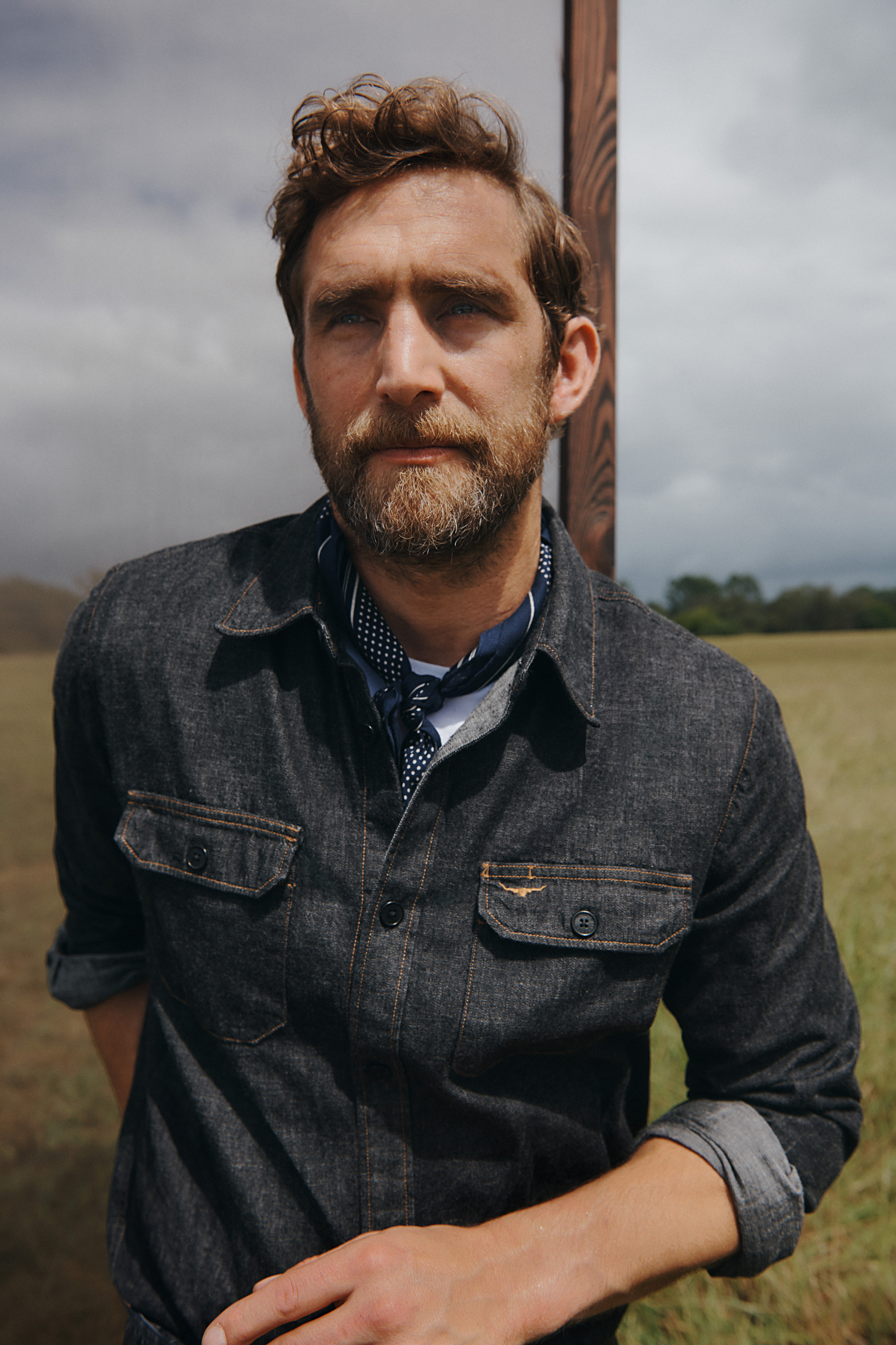 Bearded man in a denim shirt poses outdoors against a rural backdrop with a cloudy sky. Fashion shot with natural light.