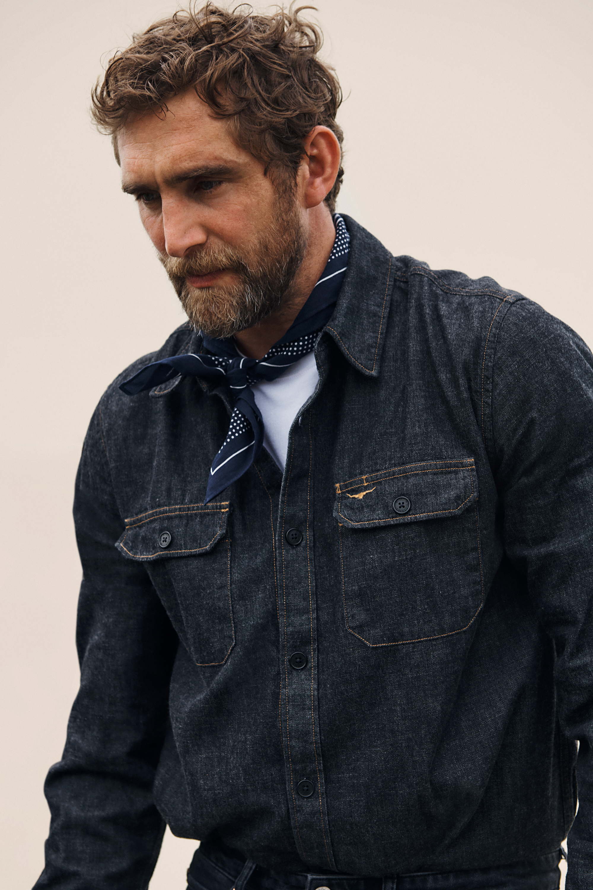 Stylish adult male with curly hair and a beard wears a blue denim jacket and patterned neckerchief looking down against a neutral background.