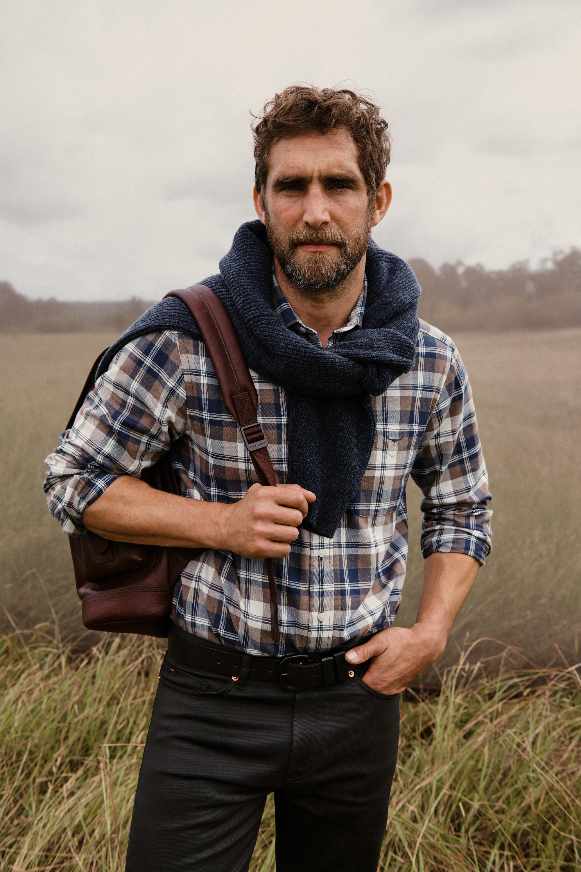 Adult male poses outdoors wearing a blue and brown plaid shirt with a navy sweater slung over his shoulders and a leather backpack.