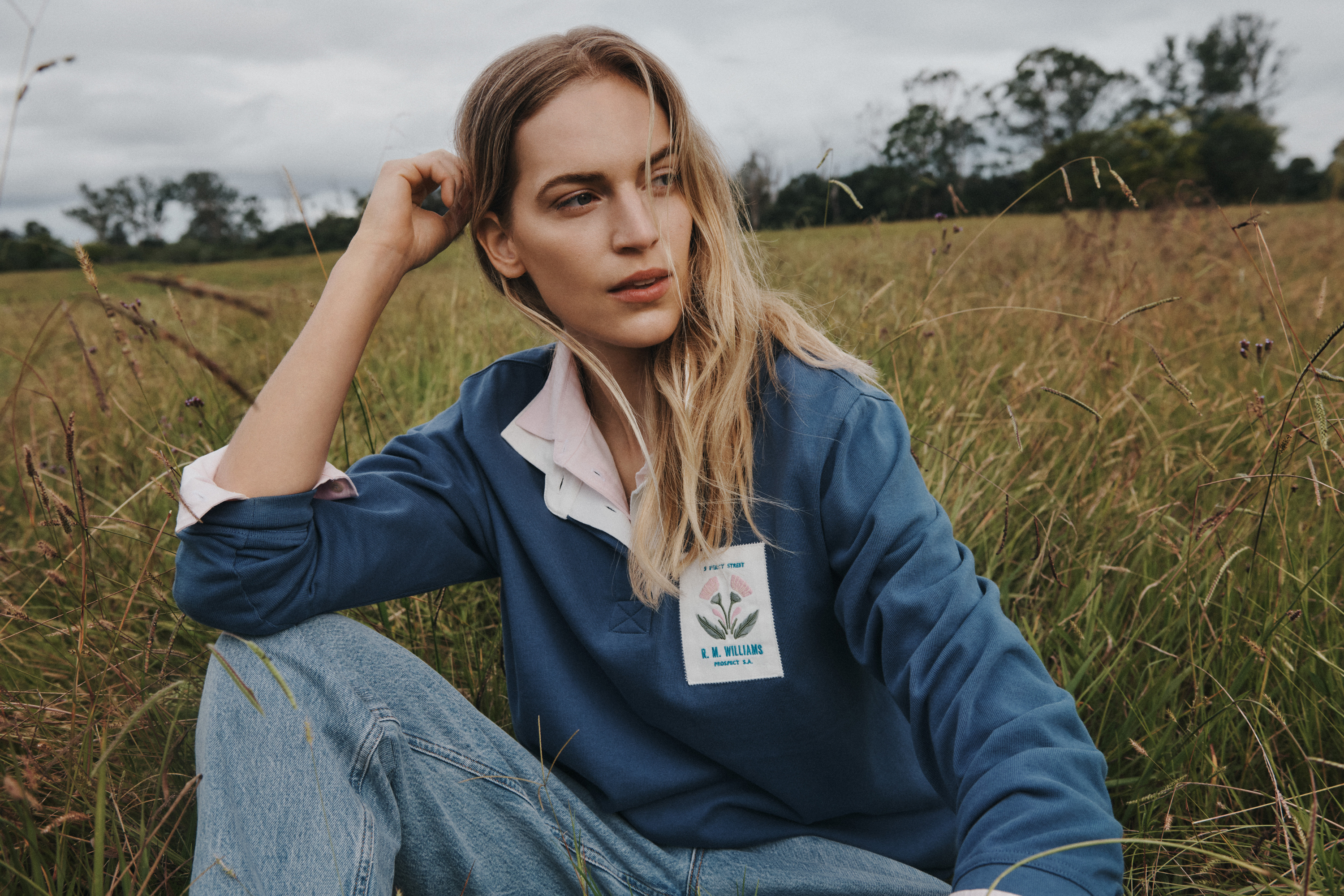 Blonde woman sits in grassy field wearing a blue long-sleeve collared shirt and jeans, posing with hand near her face.