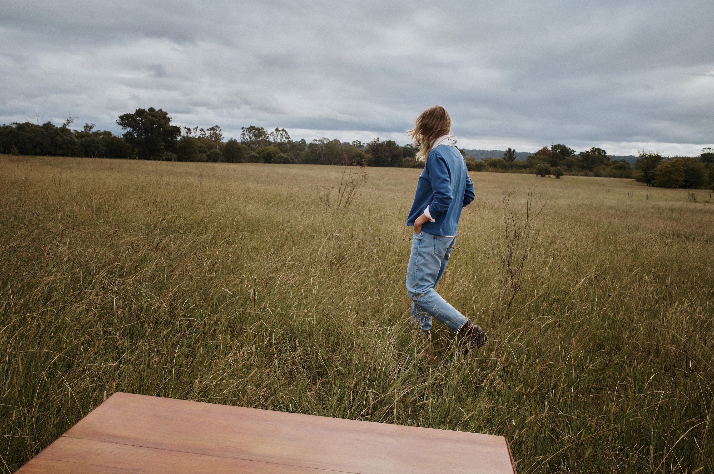 A woman with blonde hair wearing a denim jacket walks through a grassy field under an overcast sky, with a wooden surface in foreground.