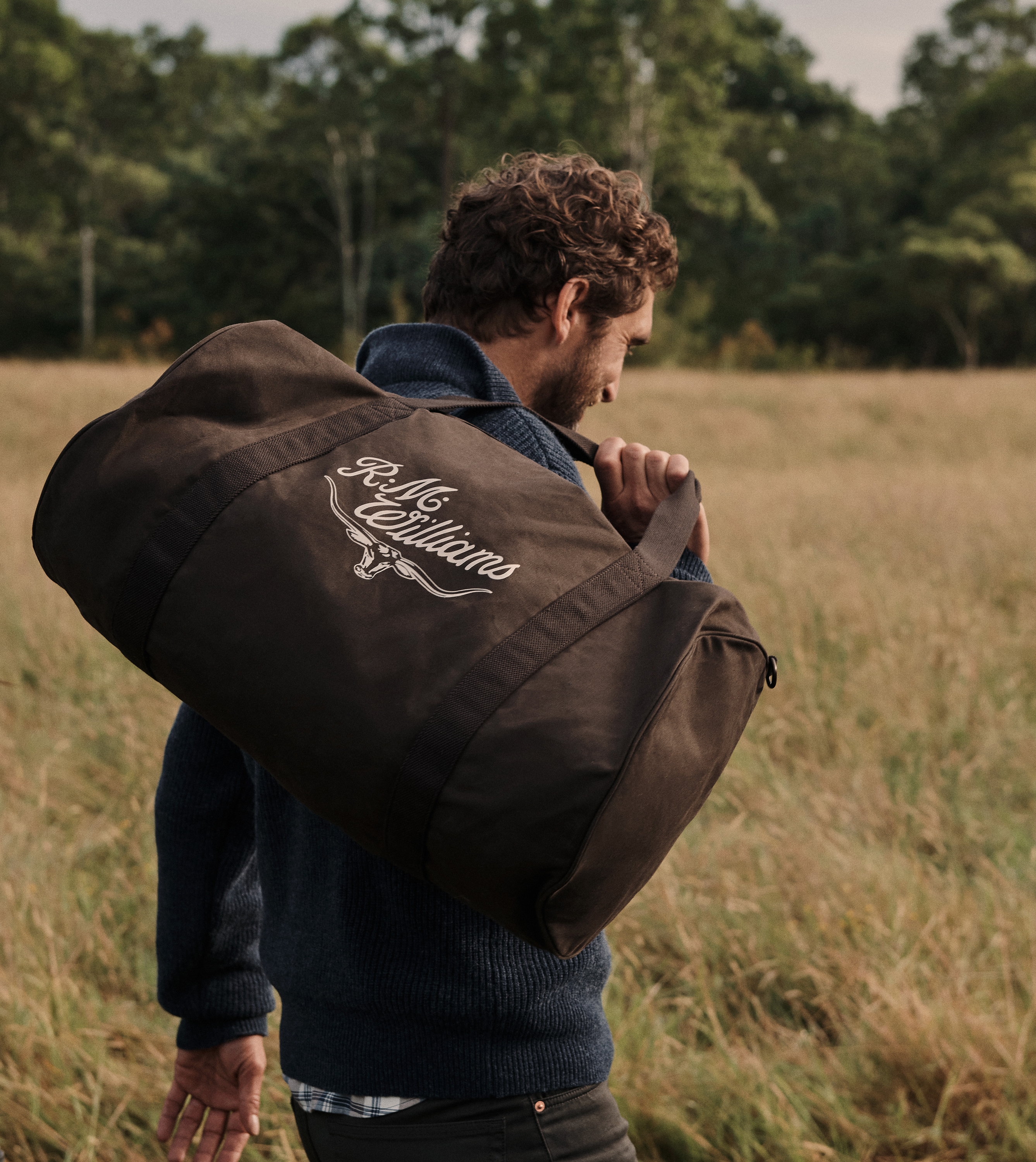 Man carries an olive-green duffel bag with the R.M. Williams logo, walking through tall dry grasses in a field, with a green treeline in the background.