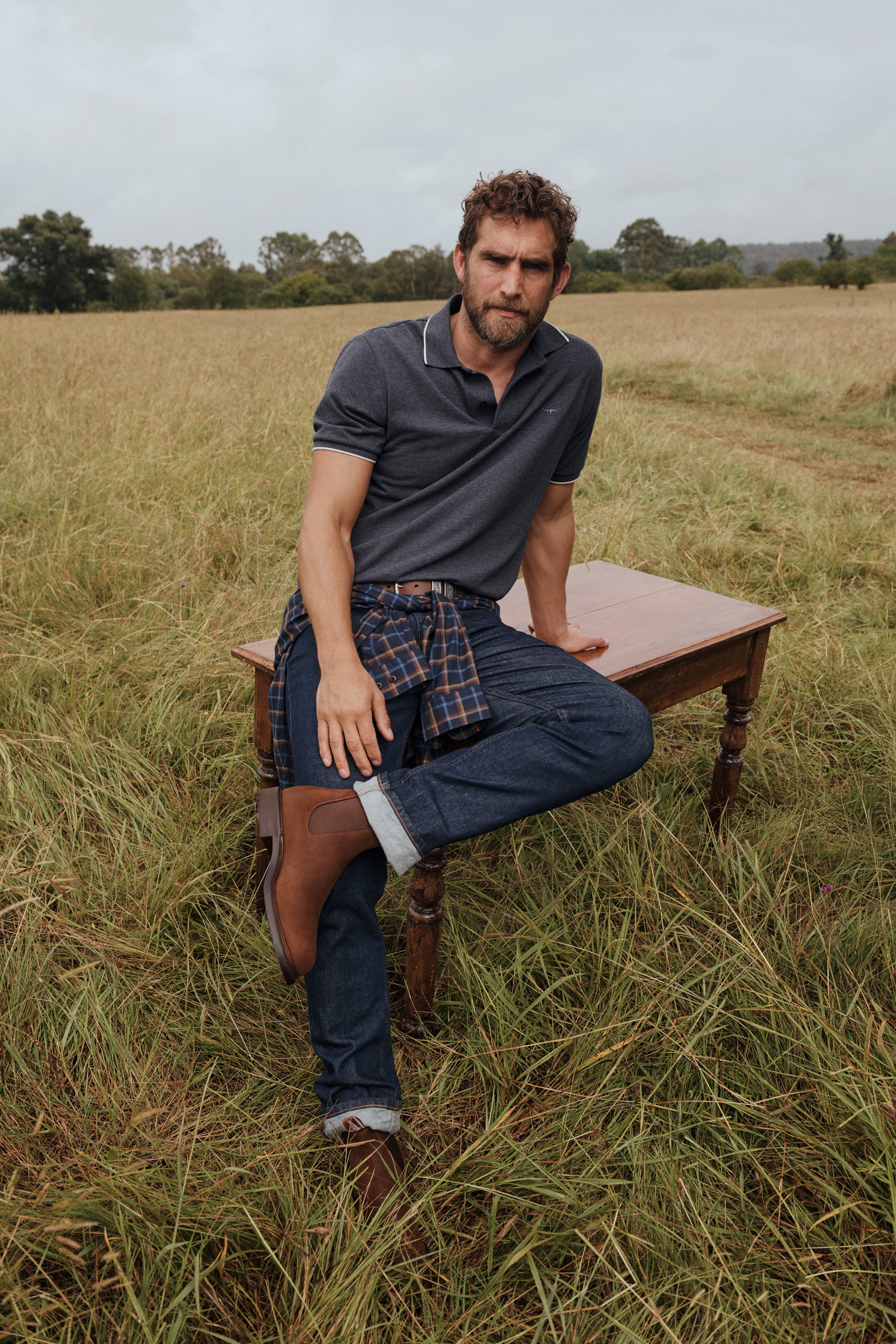 Man poses in polo shirt, jeans, and boots in a grassy field with a wooden table, wearing a plaid shirt around his waist, outdoor setting.