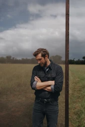 Man with arms crossed standing in a grassy field next to a vertical wooden post under an overcast sky for R.M. Williams