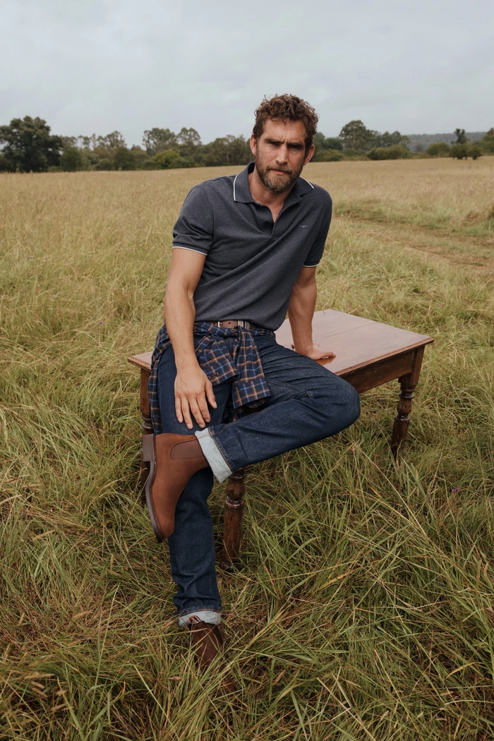 Man sitting on a wooden table in an overgrown grassy field wearing a dark polo shirt, blue jeans, and brown leather boots.