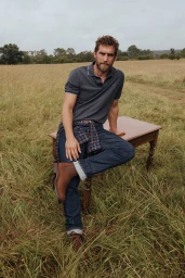 Man sitting on a wooden table in an overgrown grassy field wearing a dark polo shirt, blue jeans, and brown leather boots.