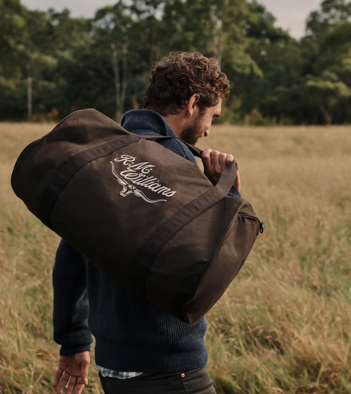 Man carrying a dark brown duffel bag over his shoulder while walking through a grassy field with trees