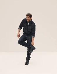 Man wearing dark denim shirt and jeans adjusting his black leather boots against a neutral background
