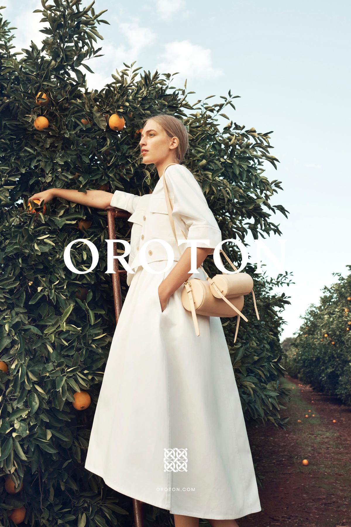 Blond model in a white dress stands next to an orange tree, holding a tan handbag in a sunny outdoor setting. Fashion advertising.