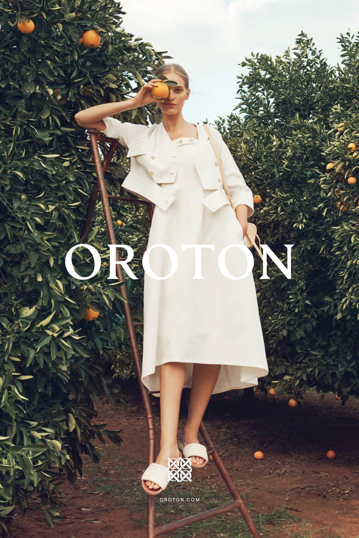 A woman in a white dress stands on a ladder in an orange grove, holding an orange, in a fashion-style outdoor shot.