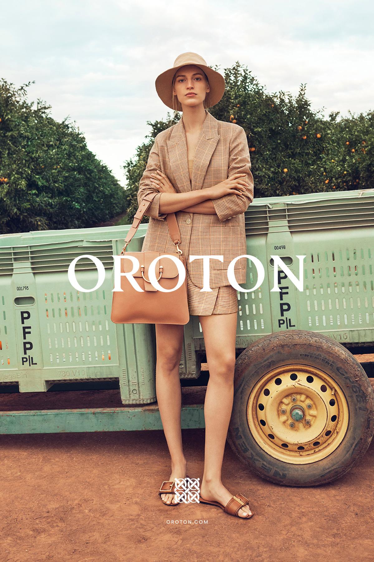 A fair-skinned woman in a beige blazer and shorts poses in front of a trailer and fruit trees wearing a hat and carrying a handbag.