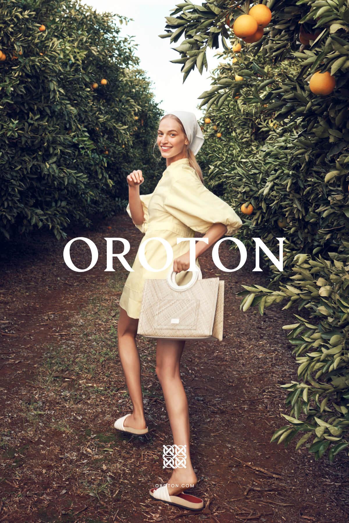 A woman in a yellow dress walks through an orange grove with a handbag, smiling slightly at the camera. Outdoor fashion shoot.