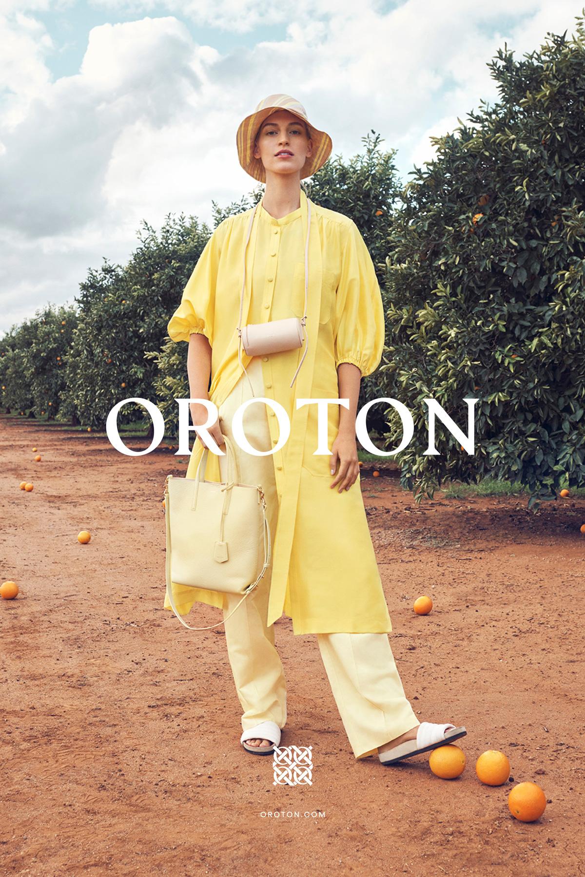 A woman in a yellow outfit poses outdoors in an orange grove with a matching handbag, styled with a hat, against a cloudy sky.