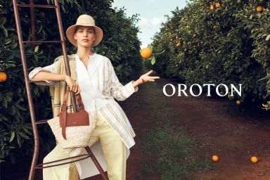 Model in a sun hat leaning against a ladder in an orange orchard holding a straw handbag