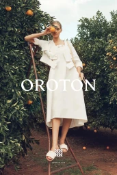 Model standing on a wooden ladder in an orange orchard holding up an orange in front of her eye, wearing a white dress and jacket.