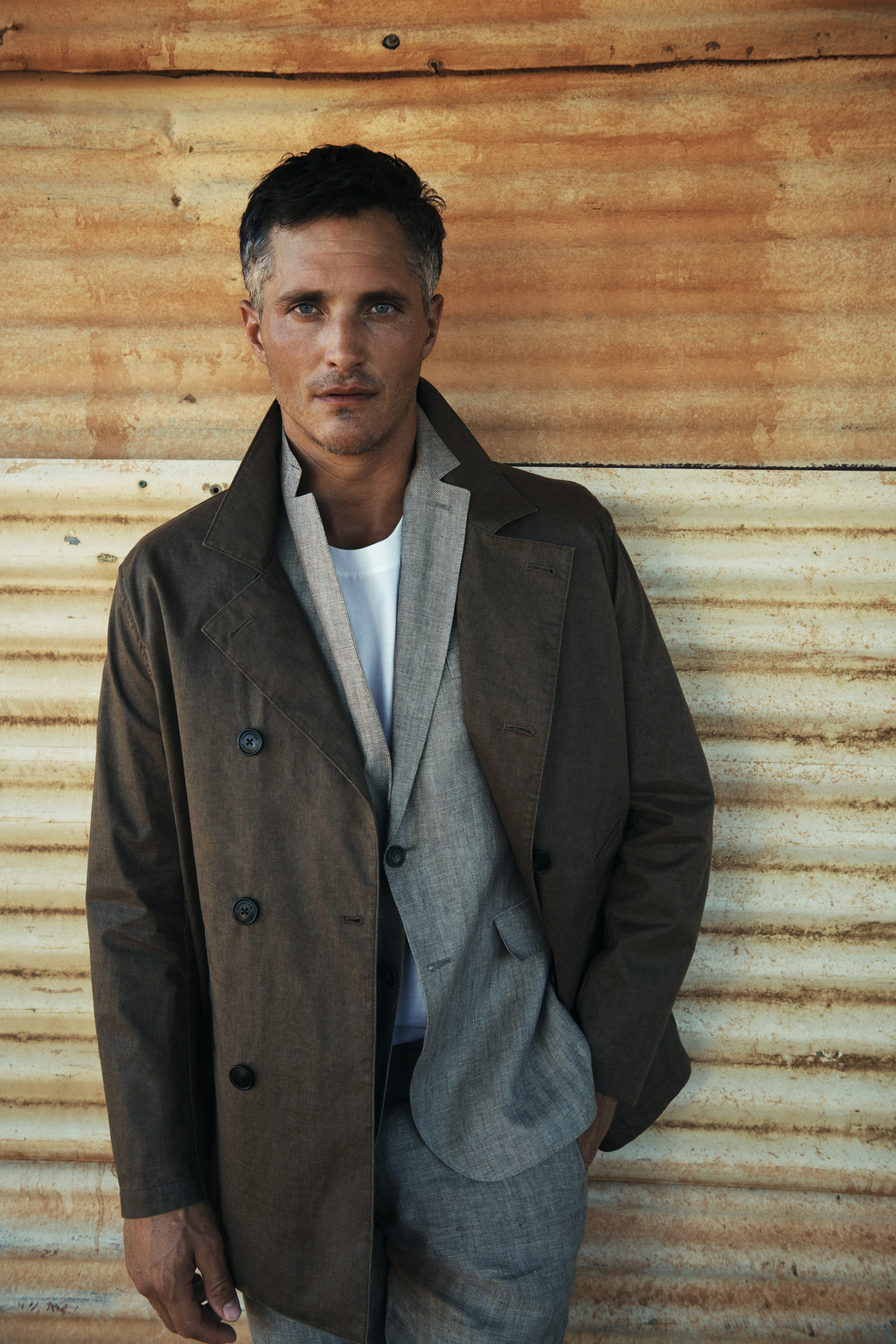 Adult male poses in a brown jacket and grey suit against a textured wall, looking directly at the camera with a neutral expression.